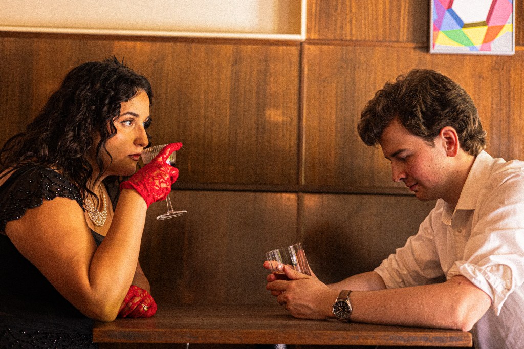 An image of Giulia and Flynn in costume as the Macbeths, at a table in a dimly lit bar, drinking together